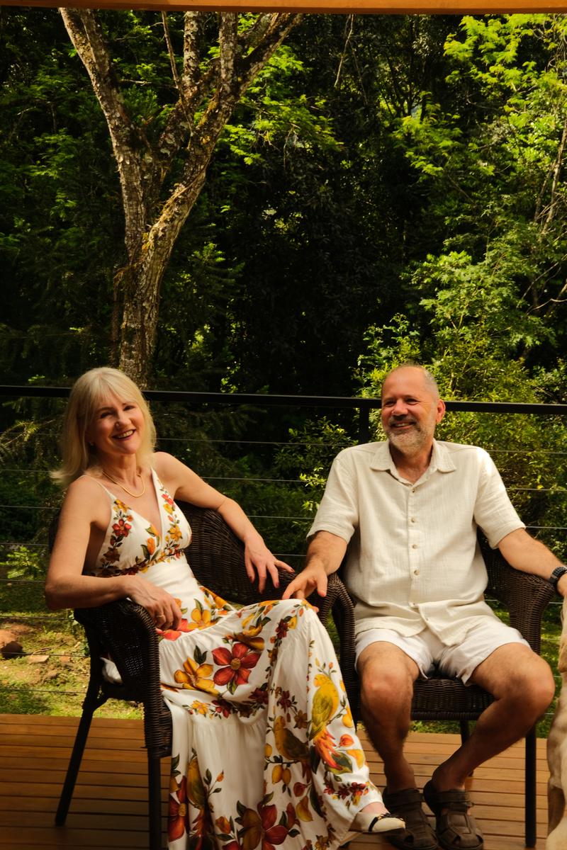 Angela e Jorge sentados lado a lado na varanda de uma das cabanas, sorrindo, com a mata ao fundo